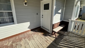 Un porche couvert avec une terrasse en bois, une porte blanche et une fenêtre munie d’une moustiquaire.