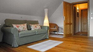 Een groene bank met bloemenkussens, een witte vloerlamp, een houten stoel en een houten deur die naar een andere kamer leidt.