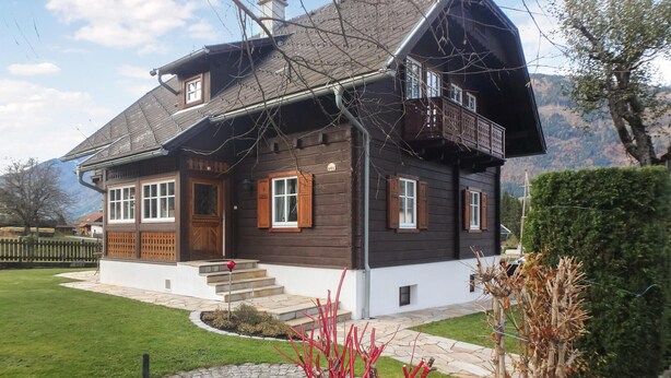 Een houten huis met twee verdiepingen, een balkon, een veranda en een pad van stenen dat naar de ingang leidt.