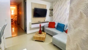 A modern living room with a flat-screen TV mounted on a wooden panel, a grey sofa with colorful cushions, a small wooden coffee table, and a patterned curtain.