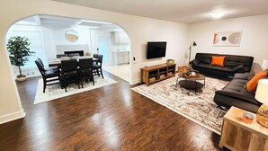 Un salon moderne avec un canapé noir, une table basse et un téléviseur. On y trouve également un coin repas avec une table et des chaises, et une cuisine à l’arrière-plan.