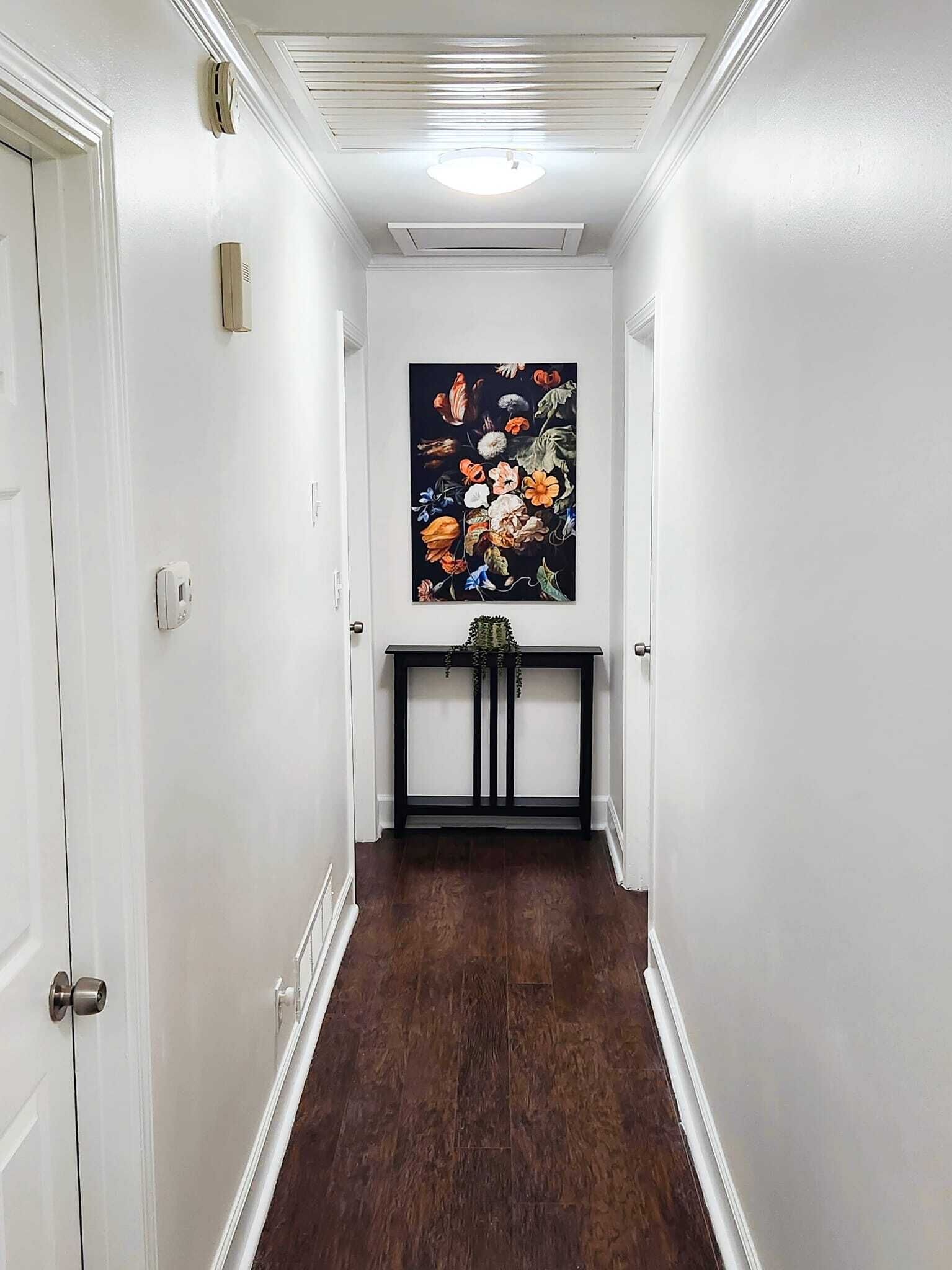 A hallway with white walls, a dark wooden floor, a ceiling light, a painting of flowers, and a small console table with a plant.