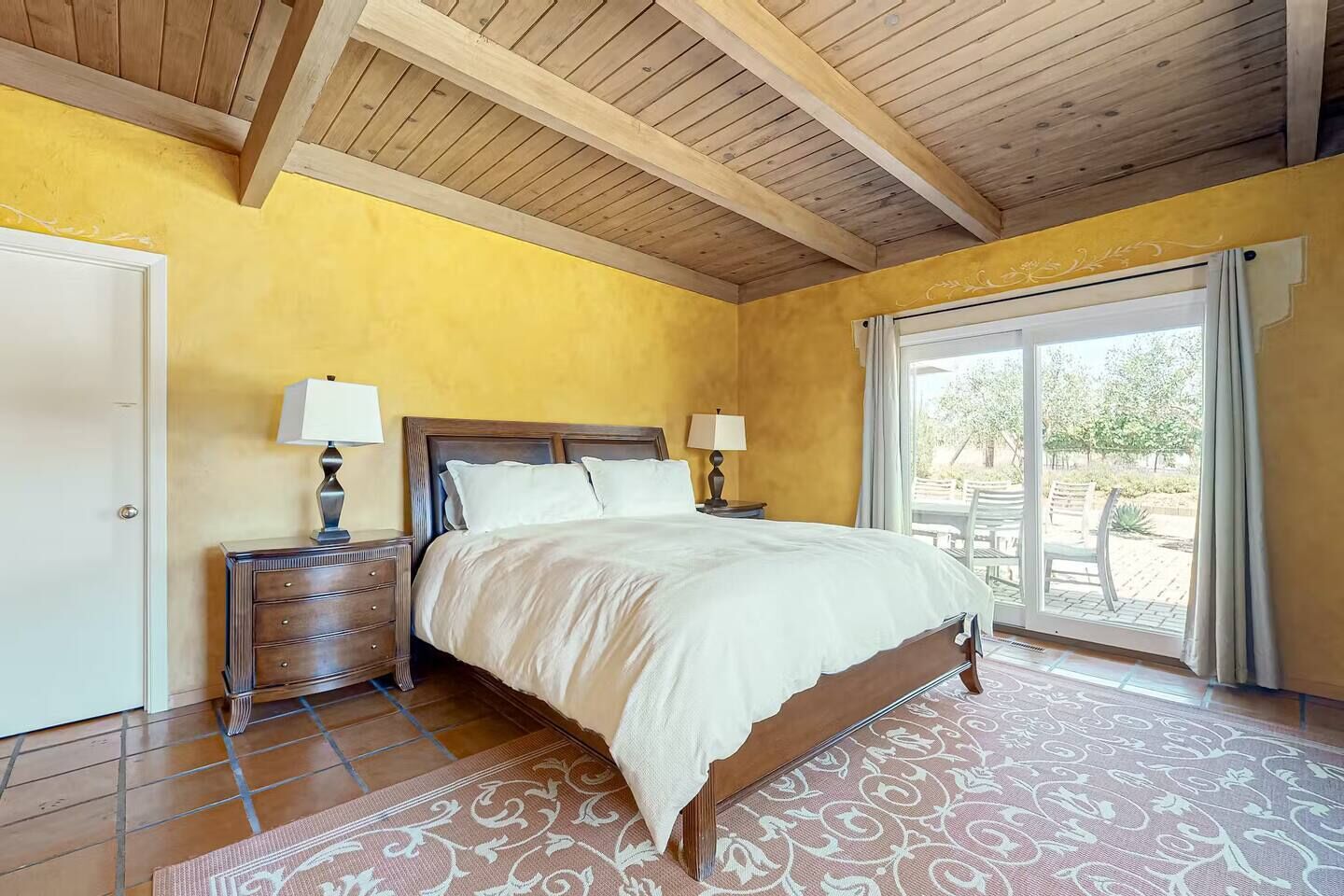 Warm & dreamy primary bedroom with golden Tuscan walls, rustic wood-beamed ceilings, a king bed dressed in crisp white linens & sliding doors to the patio.