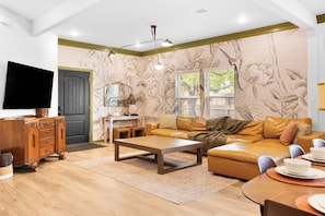 Una sala de estar con sofá, mesa de centro, aparador de madera, televisor de pantalla plana y una pared con estampado floral.