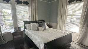 Ein Schlafzimmer mit Bett, Nachttisch, Deckenventilator und einem Fenster mit Vorhängen.