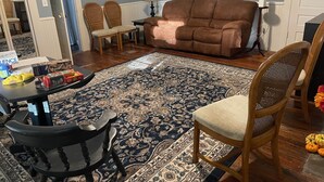 Ein Wohnzimmer mit einem braunen Sofa, zwei Korbstühlen, einem Holztisch mit Lampe und einem gemusterten Teppich.