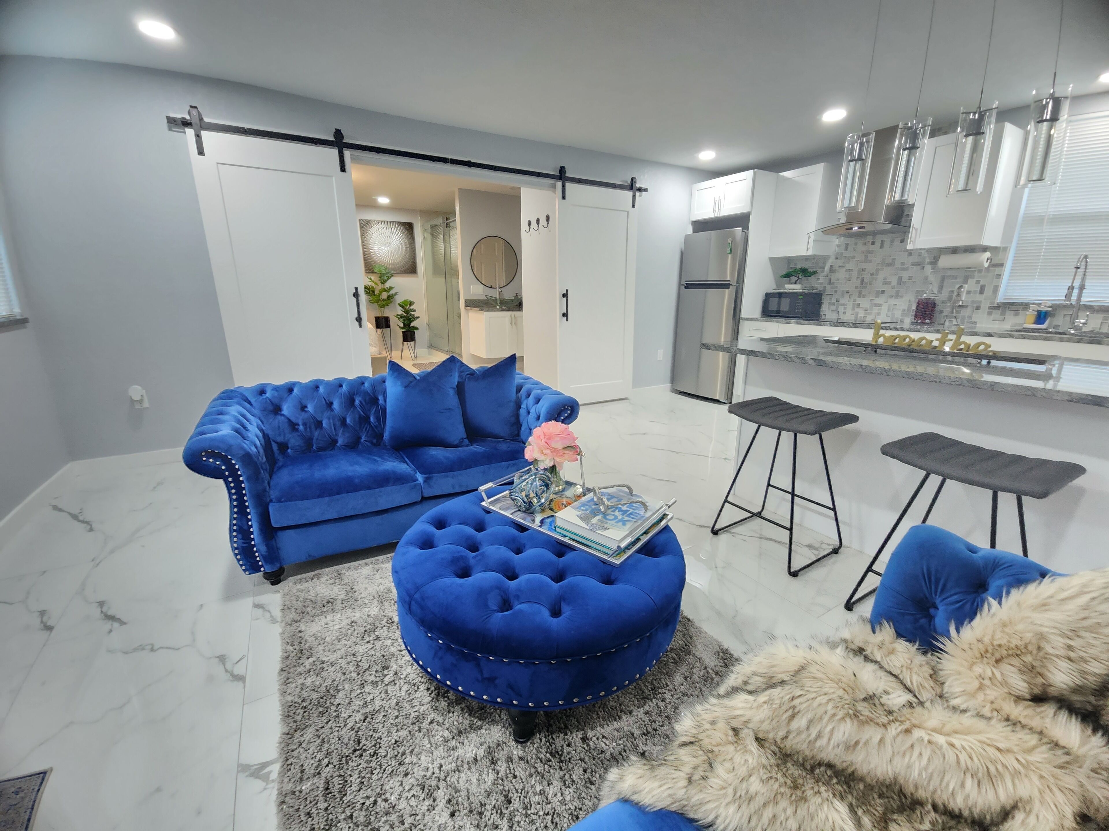 A modern living room with a blue sofa, ottoman, and matching cushions, a glass coffee table, and a fur throw.