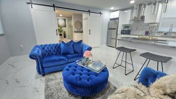 A modern living room with a blue sofa, ottoman, and matching cushions, a glass coffee table, and a fur throw.
