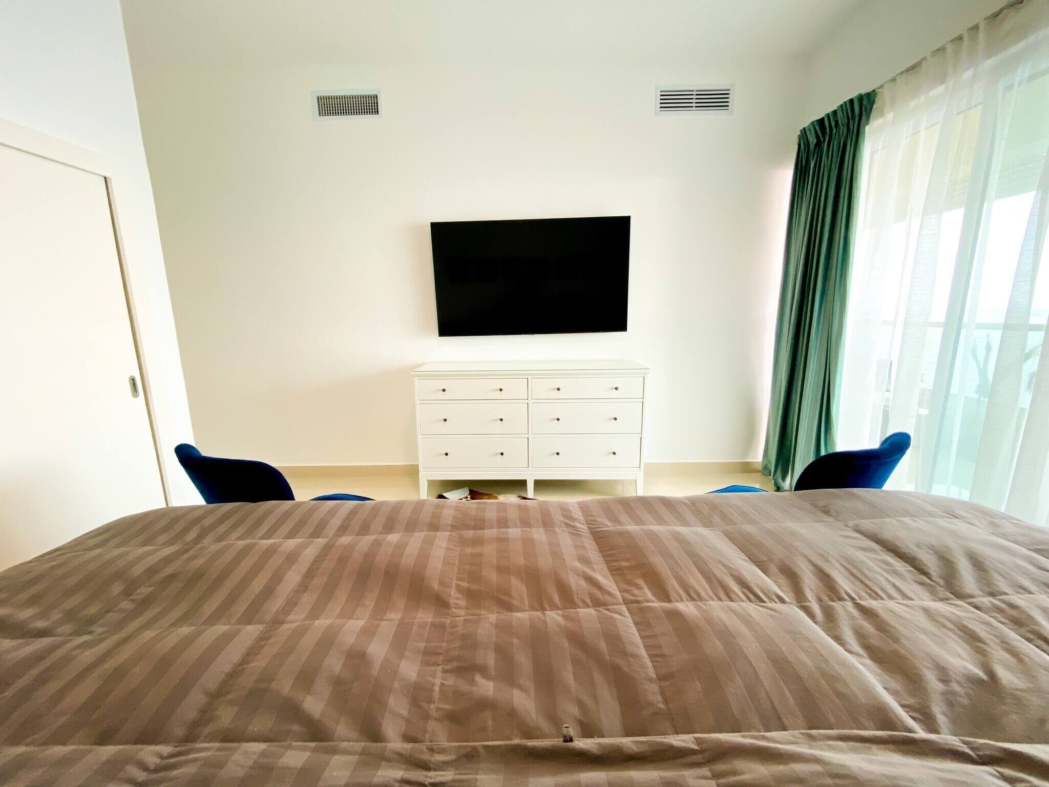 A bedroom with a bed, a white dresser, a flat-screen TV, and a window with curtains.