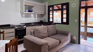 A compact kitchen with a stove, oven, and white cabinets. A beige sofa is positioned near a window with wooden shutters. - Ancoragem (Bombinhas)