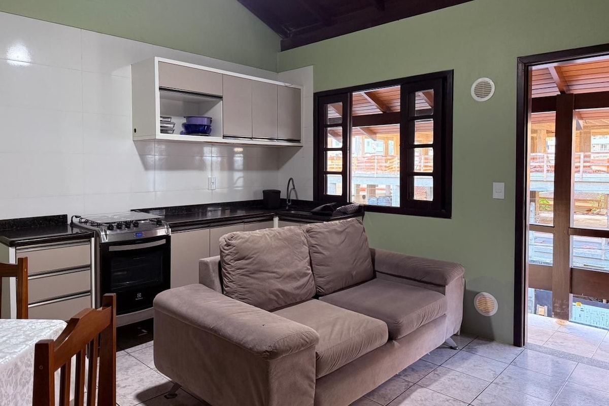 A compact kitchen with a stove, oven, and white cabinets. A beige sofa is positioned near a window with wooden shutters.