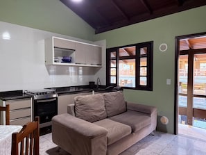 A compact kitchen with a stove, oven, and white cabinets. A beige sofa is positioned near a window with wooden shutters. - Ancoragem (Bombinhas)