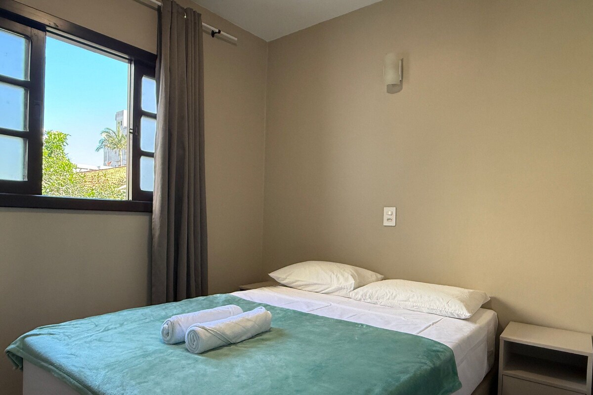 A single bed with a green blanket and two rolled towels, a nightstand with drawers, a window with curtains, and a wall-mounted light fixture.