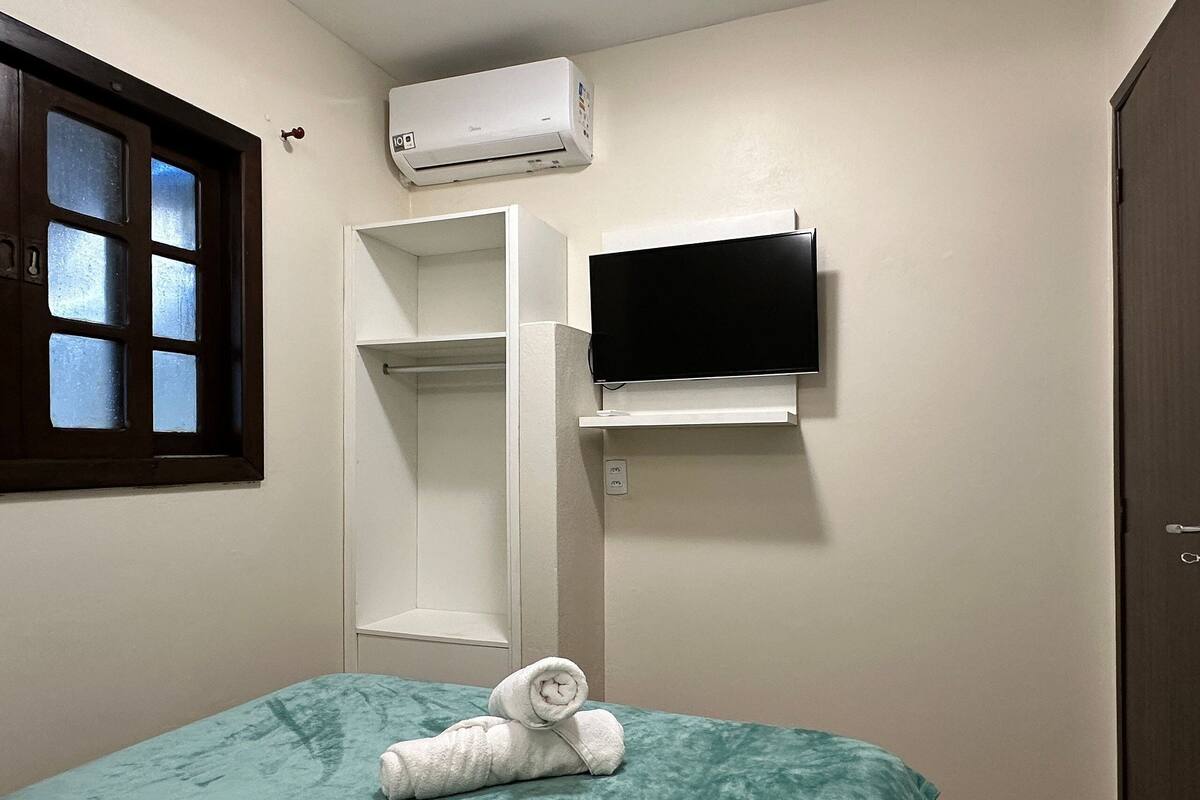 Ein kleines Einzelzimmer mit einem türkisfarbenen Bettbezug, einem zusammengerollten Handtuch auf dem Bett, einem an der Wand montierten Fernseher und einer Klimaanlage.