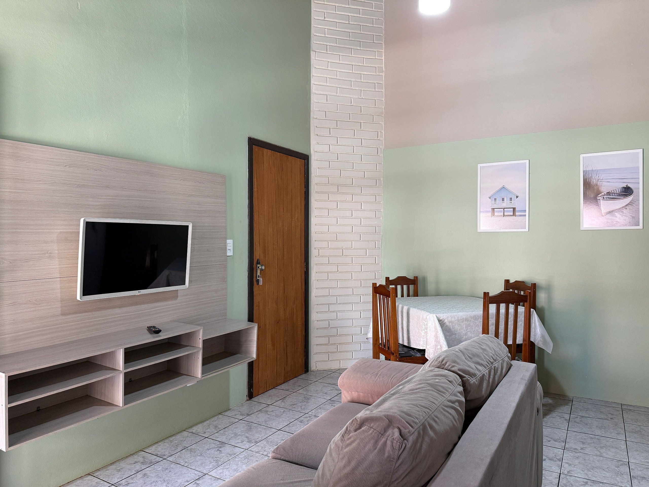 Een woonkamer met een flatscreen televisie aan een houten wand, een eettafel met stoelen en een bank.