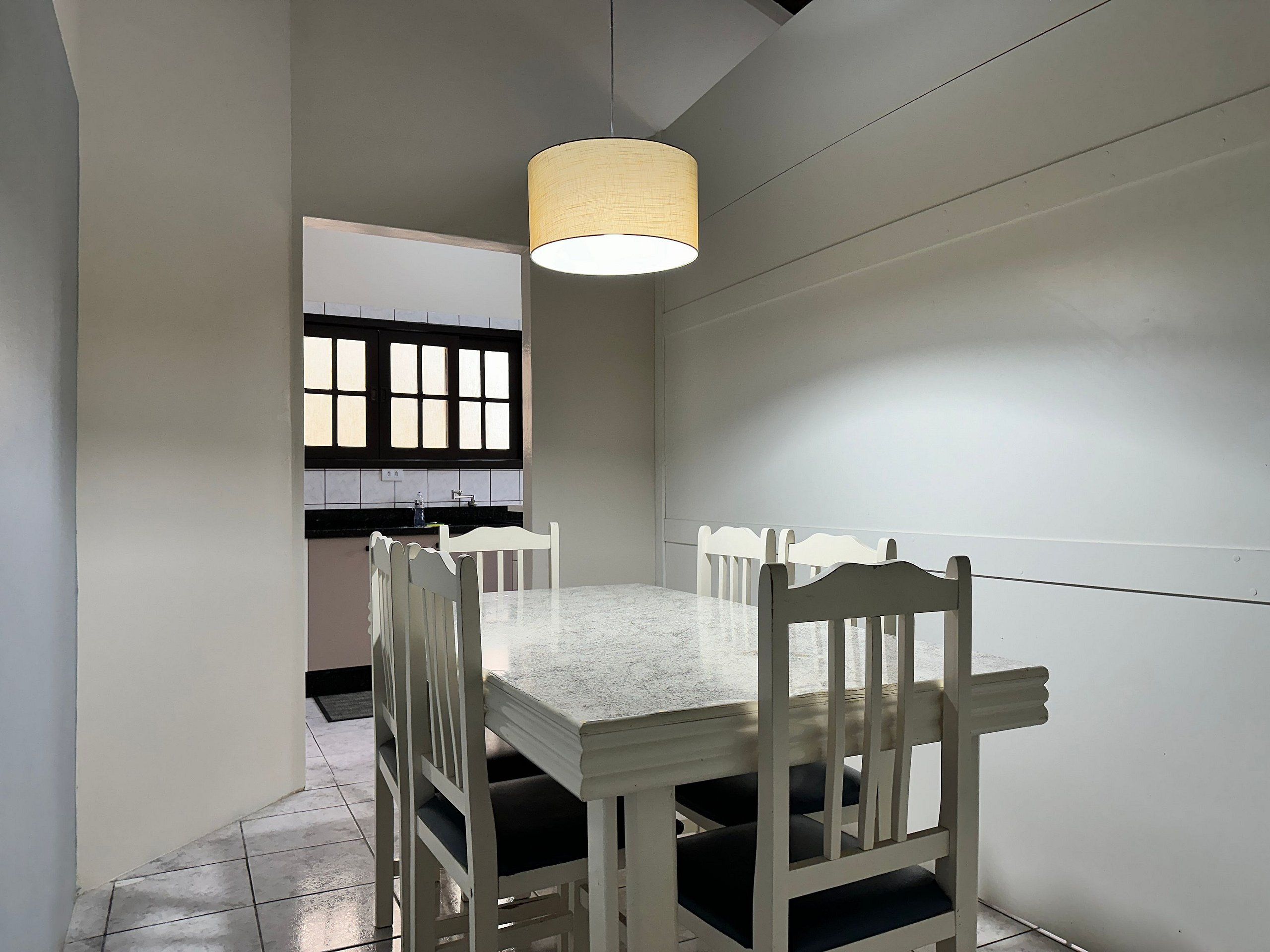 A dining area with a white table and chairs, a hanging light fixture, and a window with blinds.