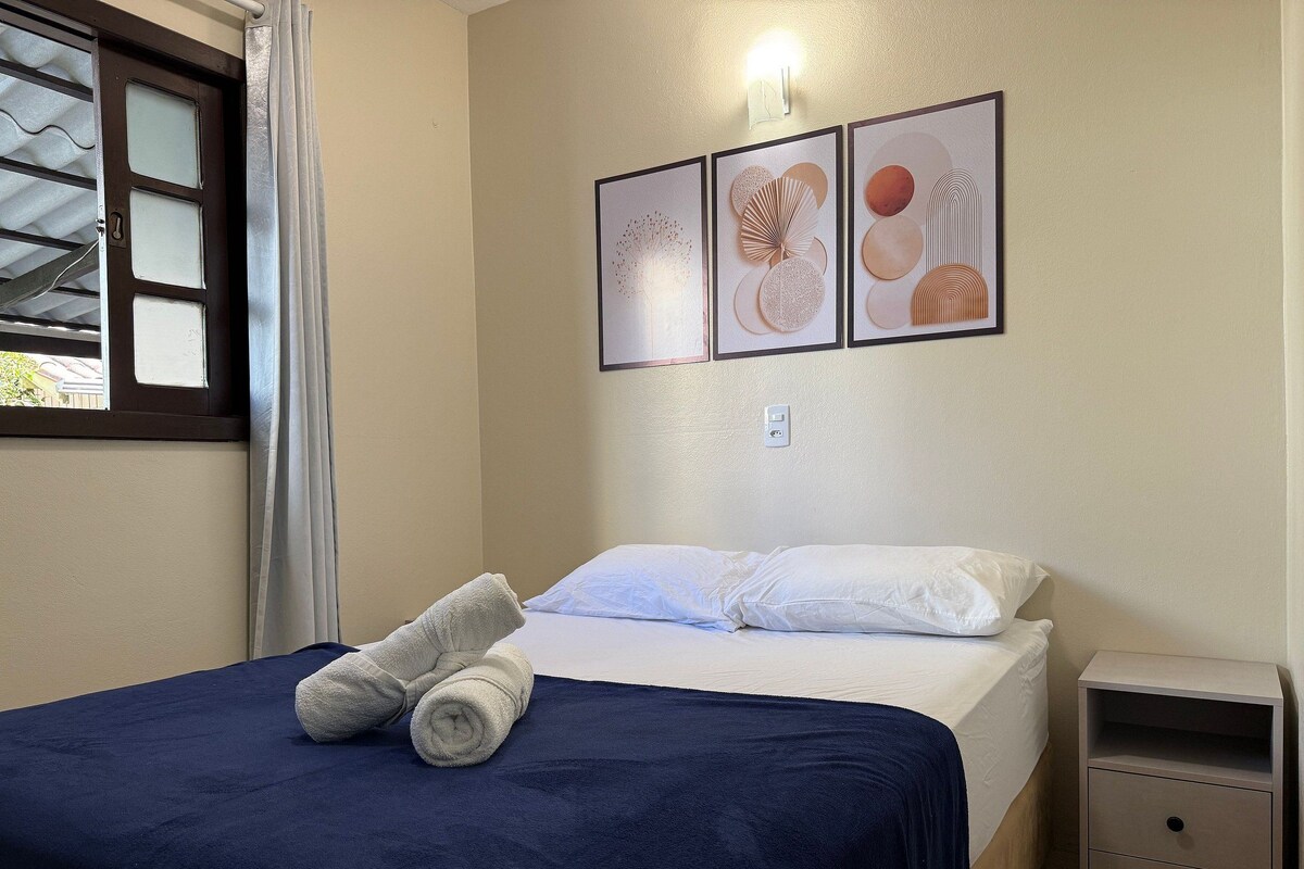 A neatly made bed with a blue bedspread and two rolled towels, a small bedside table with drawers, and three framed artworks on the wall.