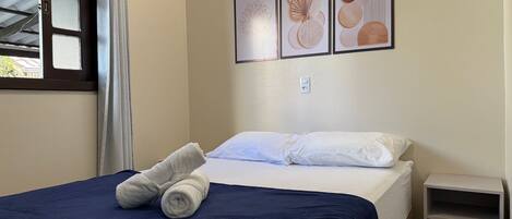 Una cama bien hecha con una colcha azul y dos toallas enrolladas, una mesita de noche con cajones, y tres obras de arte enmarcadas en la pared.