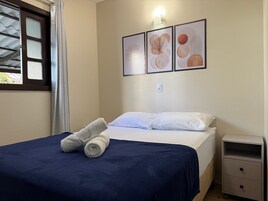 Una cama bien hecha con una colcha azul y dos toallas enrolladas, una mesita de noche con cajones, y tres obras de arte enmarcadas en la pared.