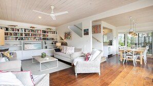 A spacious living area with a large bookshelf, a white sofa, a wicker chair, and a dining area with a wooden table and chairs. - Bush setting and easy walk to beach (KILLCARE)