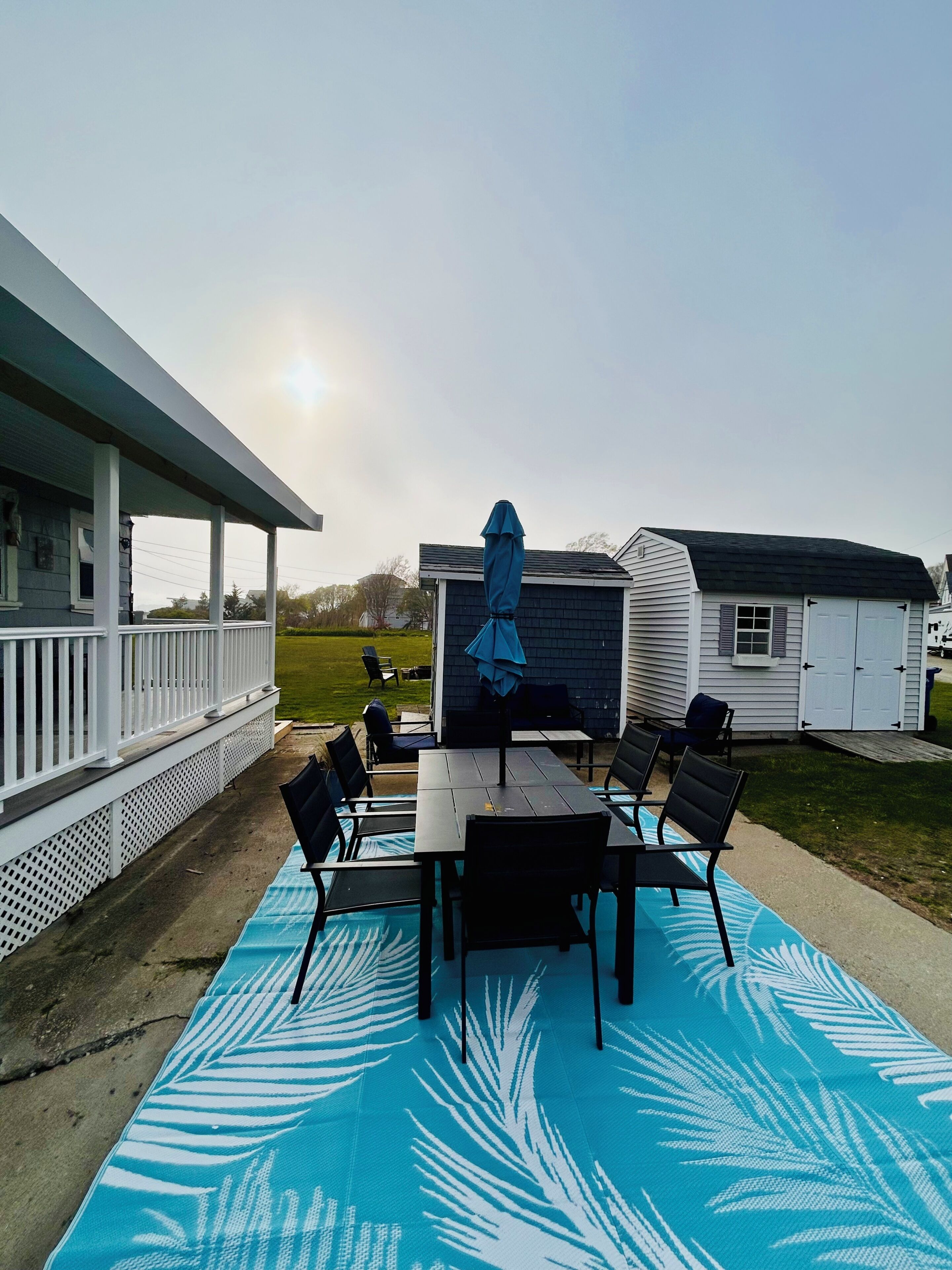 Une terrasse couverte avec une table et des chaises, un parasol bleu et un petit abri de jardin en arrière-plan.