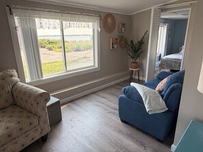 A living room with a large window, a blue sofa, a patterned armchair, and a small side table with a plant. - Beach Lovers Paradise!  Cozy 3 bedroom home steps from the beach.  (Fairhaven)