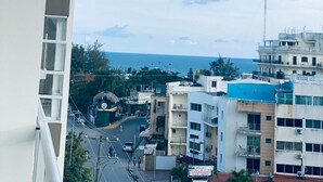 A balcony overlooking a street with buildings, a clear sky, and a distant view of the sea. - Apartamento Acogedor Cerca de la Playa (Boca Chica)