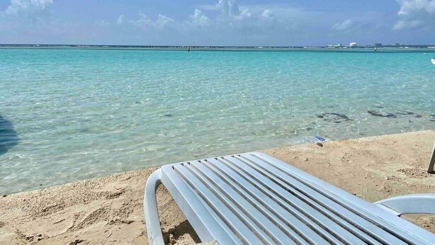A beach with clear turquoise water, a sandy shore, and a metal lounge chair. - Apartamento Acogedor Cerca de la Playa (Boca Chica)