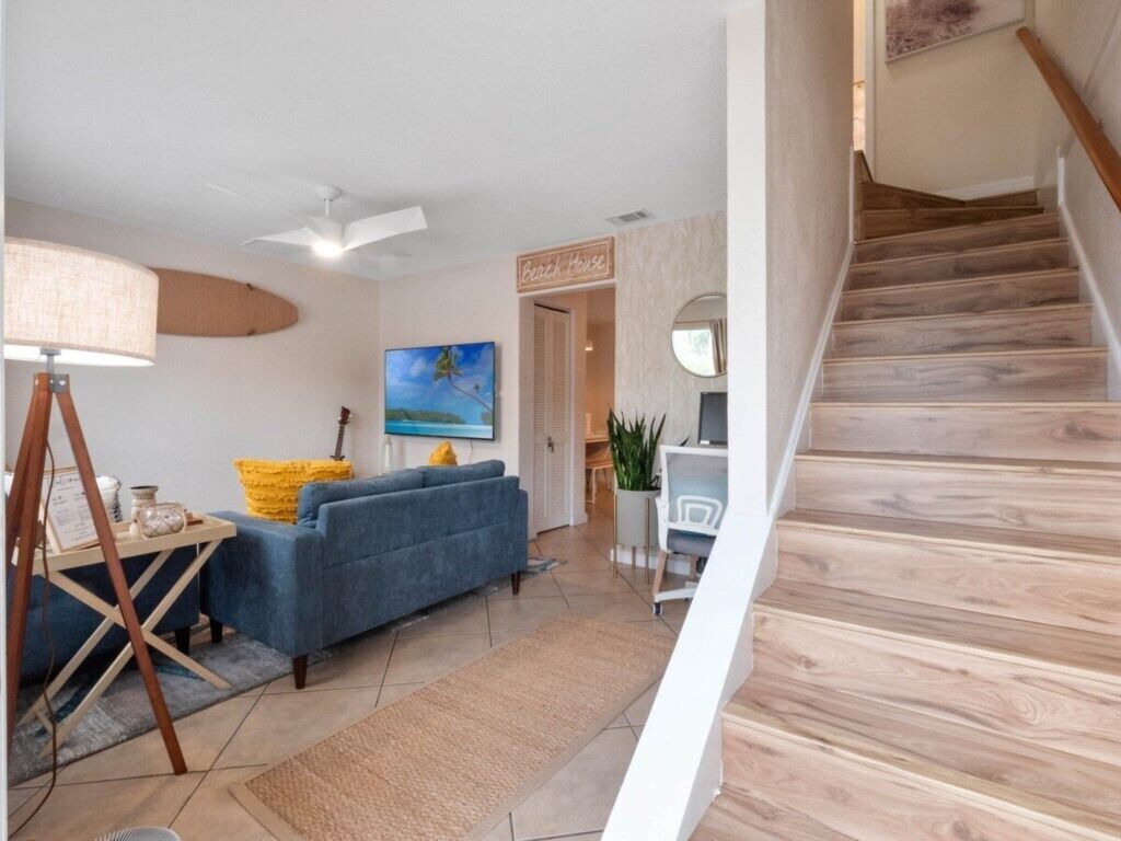 Ein Wohnzimmer mit einem blauen Sofa, einer Lampe und einem Gemälde. Eine Treppe mit Holzsitzen ist sichtbar.