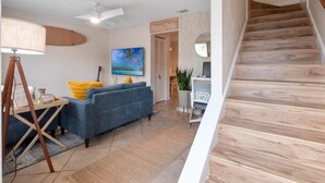 Una sala de estar con un sofá azul, una lámpara y un cuadro. Se ve una escalera con escalones de madera.