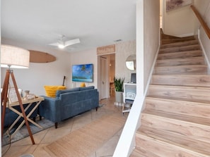Ein Wohnzimmer mit einem blauen Sofa, einer Lampe und einem Gemälde. Eine Treppe mit Holzsitzen ist sichtbar.