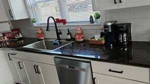 Eine Küche mit weißen Schränken, einer schwarzen Arbeitsplatte, einem Edelstahl-Geschirrspüler, einer Kaffeemaschine und einem Fenster mit Jalousien.