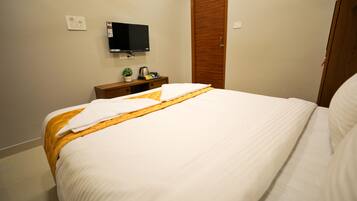 A hotel room with a neatly made bed, a television mounted on the wall, a wooden wardrobe, and a small table with a plant and a kettle.