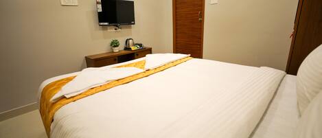 Habitación de hotel con una cama bien hecha, un televisor montado en la pared, un armario de madera y una mesita con una planta y una tetera.