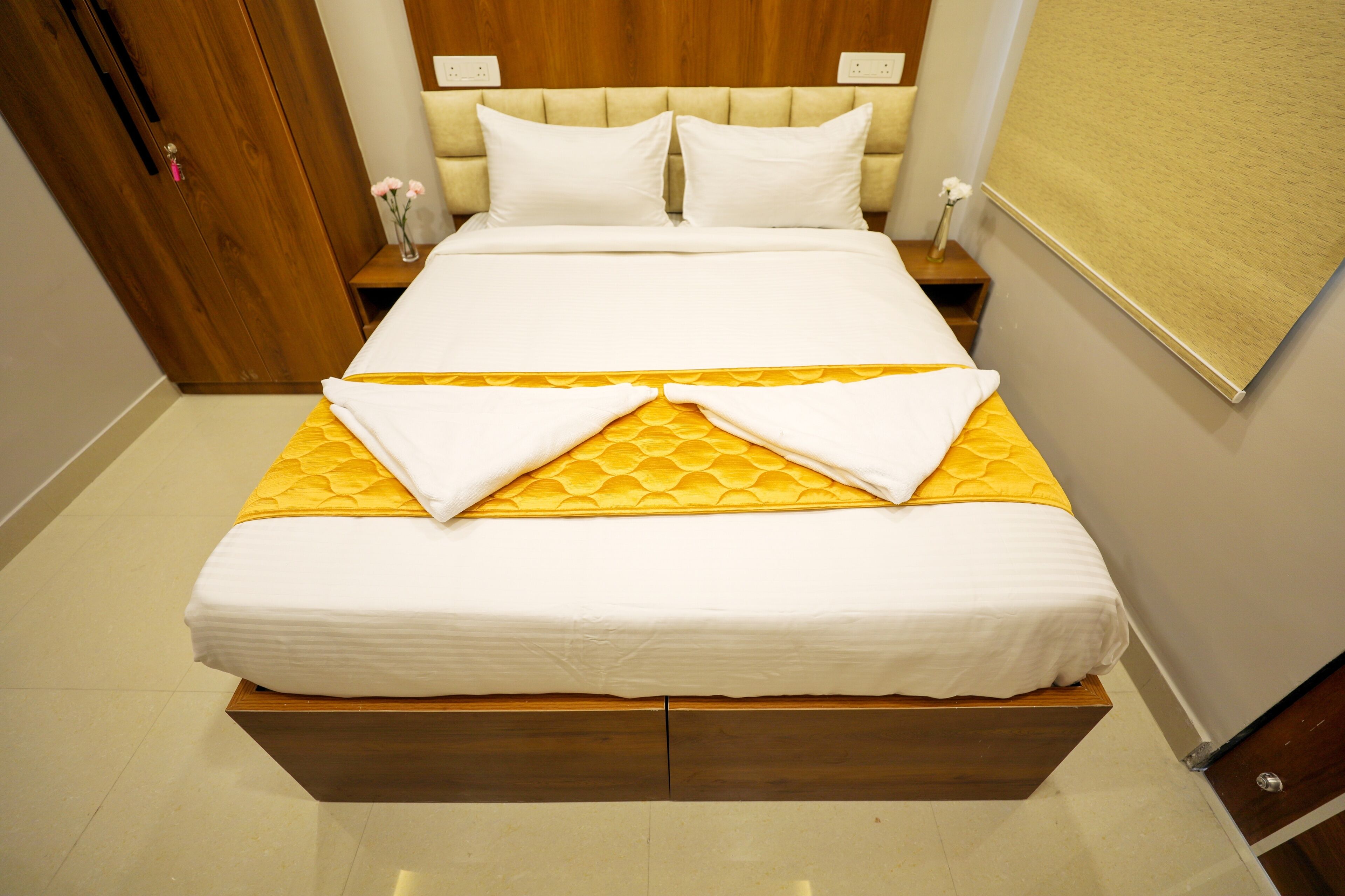 A neatly made bed with a yellow mattress cover and white linens, a wooden headboard, and a wooden nightstand with a vase of flowers.