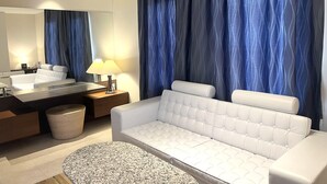 A modern hotel room with a white sofa, a round coffee table, a large mirror, a vanity with a sink, and blue curtains.