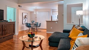 A living room with a glass coffee table, a dark grey sofa with yellow cushions, a wooden cabinet, and a dining area with a blue table and chairs. - Welcome to Your Vacation Home (Clearwater)