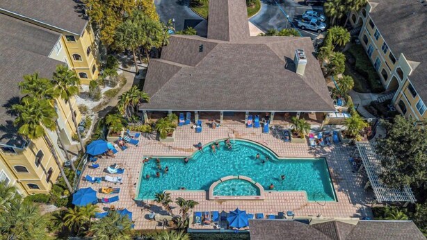 An aerial view of a hotel with a central swimming pool surrounded by lounge chairs and umbrellas, palm trees, and a parking area. - Welcome to Your Vacation Home (Clearwater)