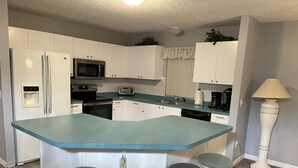 A kitchen with white cabinets, a blue countertop, a microwave, a toaster, a sink, and a coffee maker. There are three stools with green cushions.