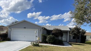 A single-story house with a garage, a front yard with grass and shrubs, and a clear blue sky with clouds.