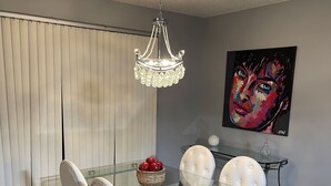 A dining area with a glass table, white upholstered chairs, a chandelier, and a wall-mounted painting.