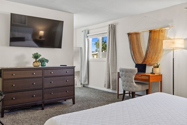 A bedroom with a bed, a dresser, a desk with a lamp, and a TV on the wall.
