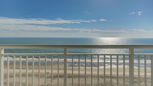 Vista da praia a partir de uma varanda com guarda-corpo, oceano e céu claro.