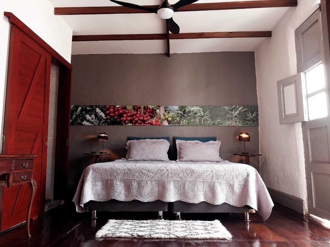 A bedroom with a bed, wooden headboard, ceiling fan, two bedside lamps, and a decorative wall hanging.