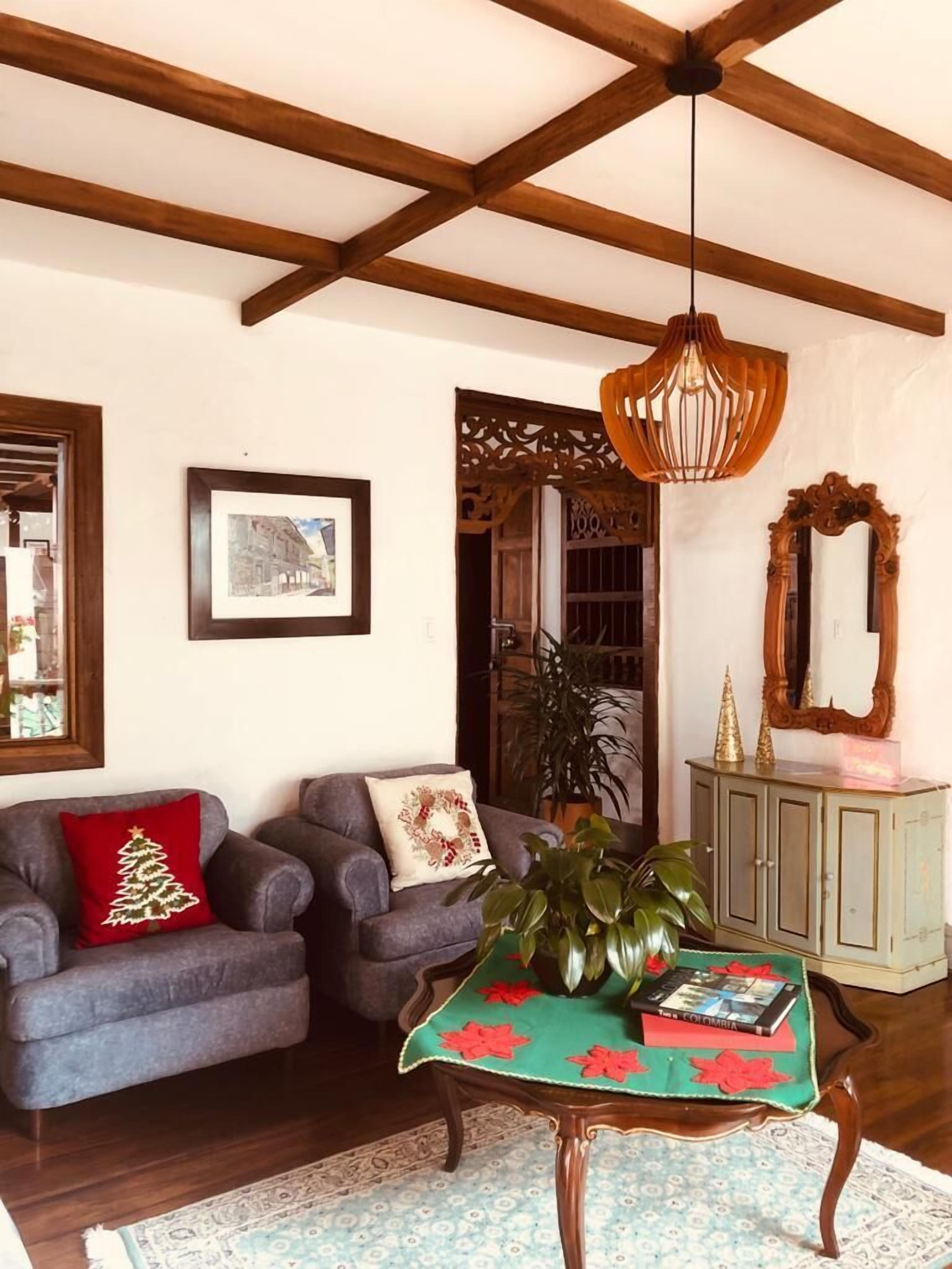 Un soggiorno con soffitto alto, travi in legno, un lampadario, un divano con un cuscino a tema natalizio, un tavolo con un libro e un mobile contenente oggetti decorativi.