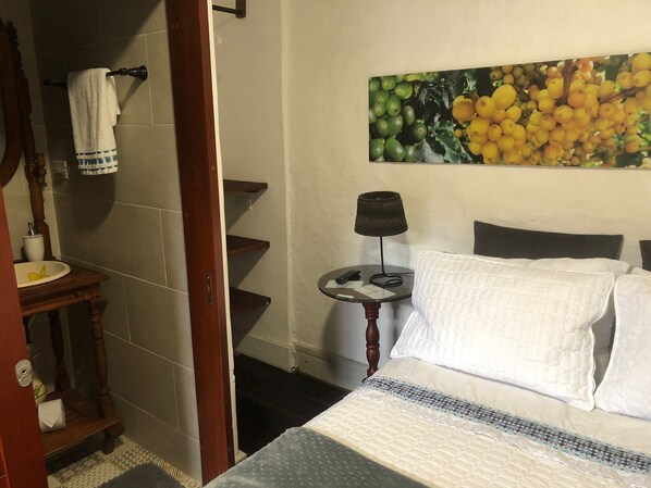 A hotel room with a bed, a bedside table with a lamp, a painting of fruit on the wall, and a bathroom with a towel rack and sink.