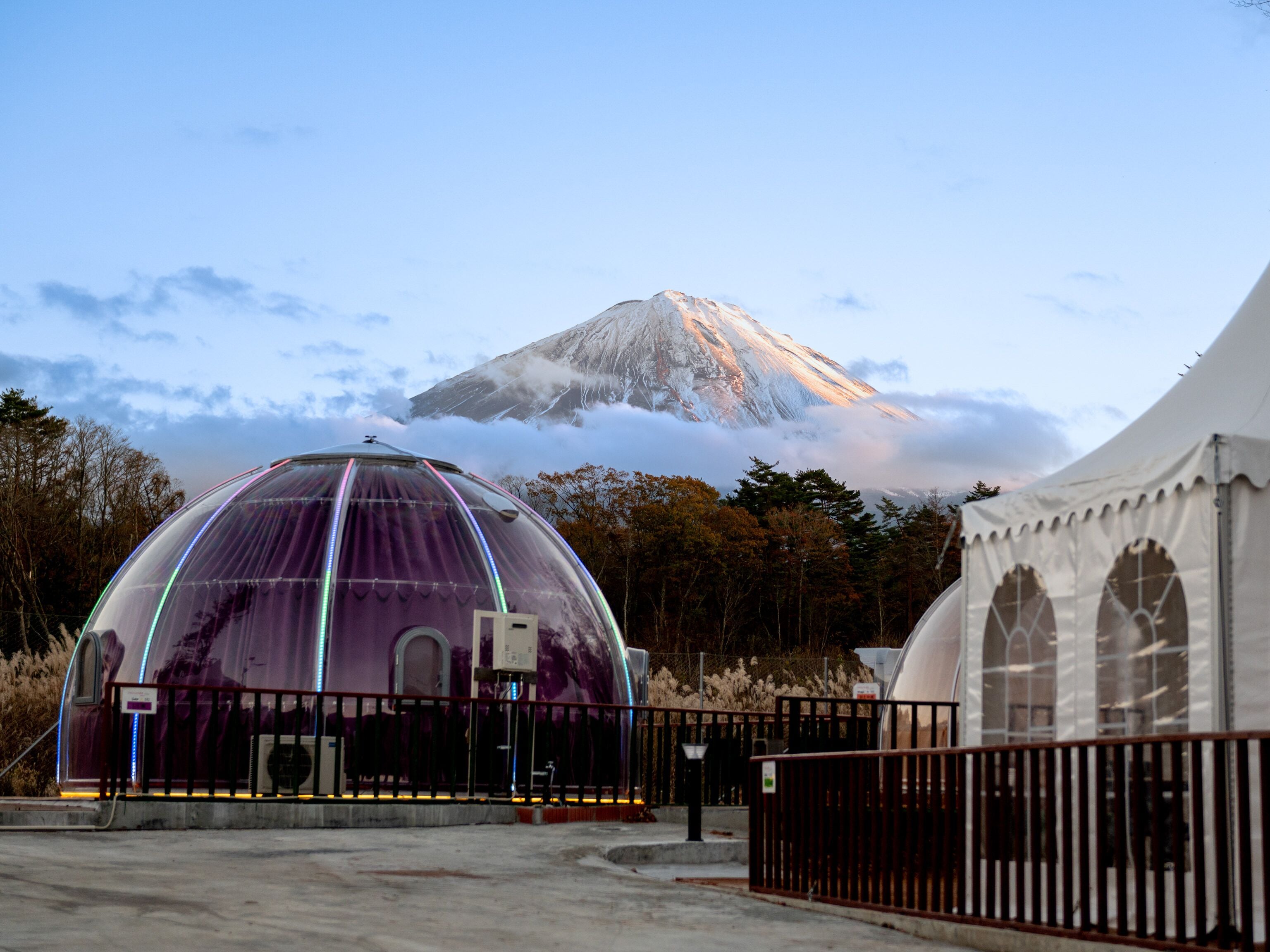 A dome-shaped building with a transparent roof, surrounded by a fence, and a white tent-like structure in the background.