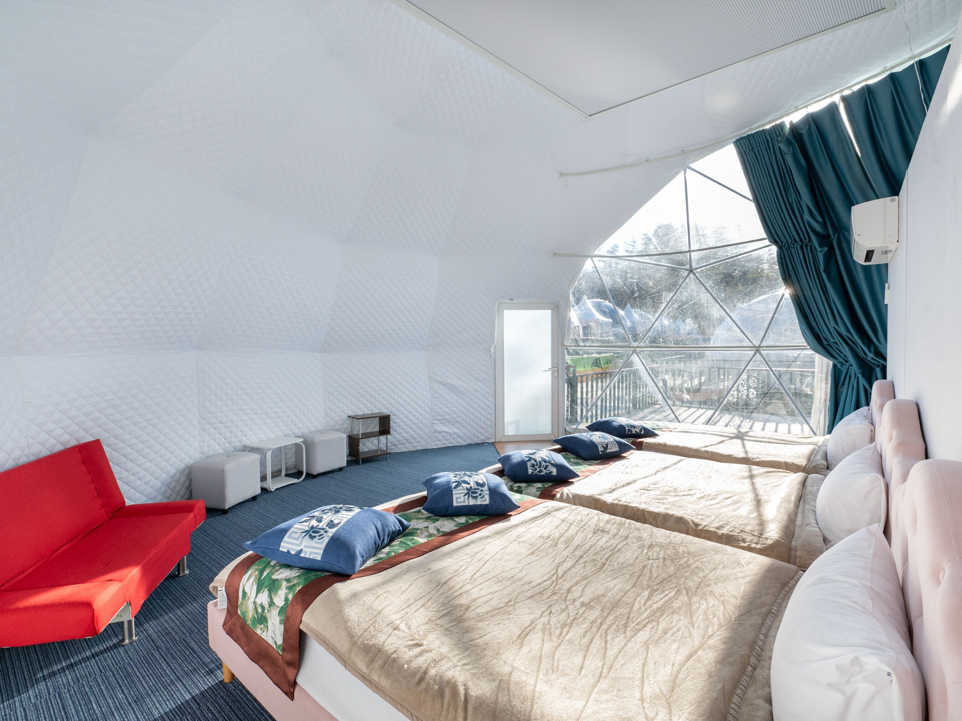 Ein Zimmer mit zwei Betten, einem roten Sessel und einem großen Fenster mit Vorhängen.