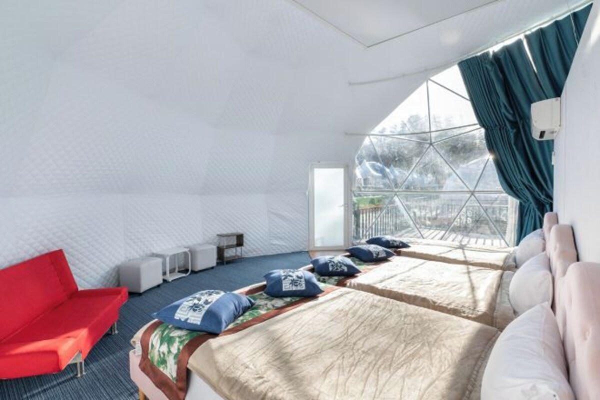 A modern, well-lit room with two beds, a red chair, and a large window with curtains.
