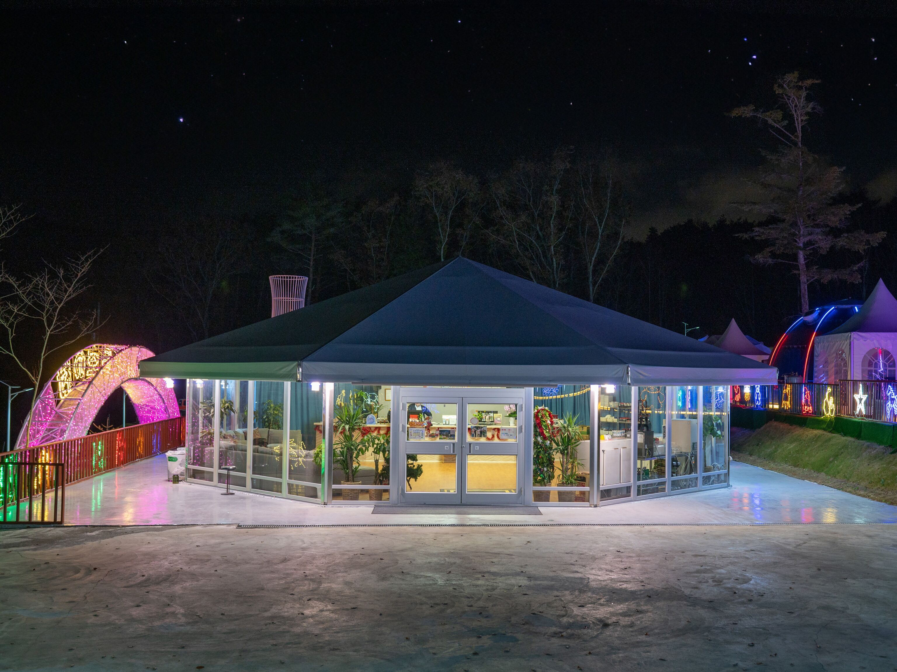 A glass-roofed pavilion with a green roof, surrounded by colorful lights and a small garden area.